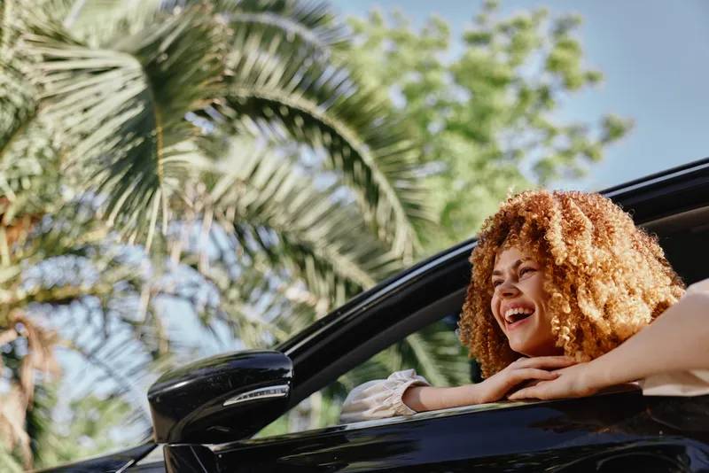 Smiling woman with curly hair leaning out of car window on a sunny day, surrounded by green palm trees and clear blue sky, showing joy and freedom.