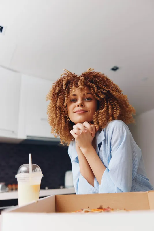 Кудрявая женщина наслаждается пиццей и напитком, сидя перед коробкой на столе