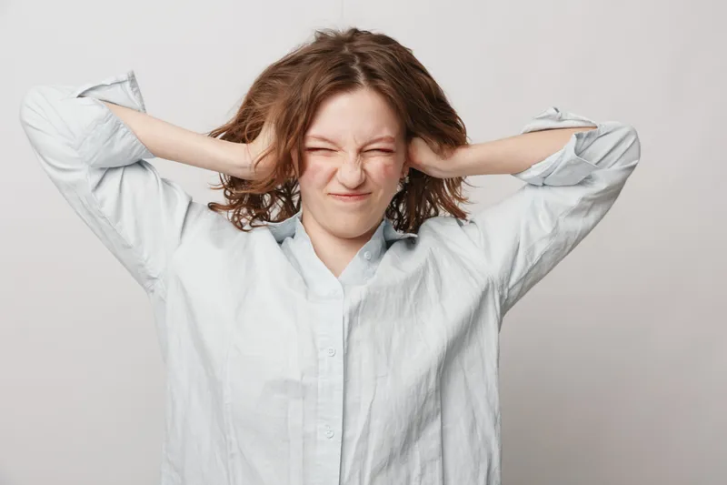 Young woman in white shirt covers ears with hands and squints eyes, expressing discomfort or stress. Isolated on light background. Concept of noise, frustration, and emotional tension.