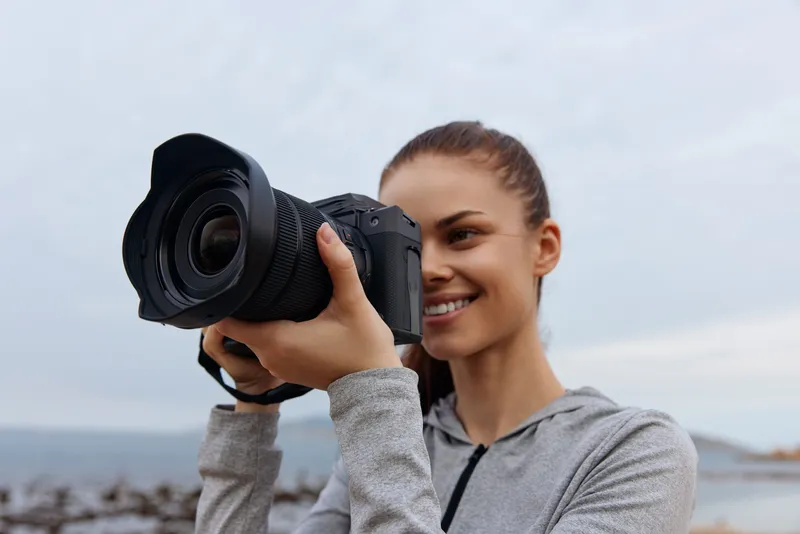 Smiling young woman with a camera capturing moments on a cloudy beach, embodying creativity and passion for photography