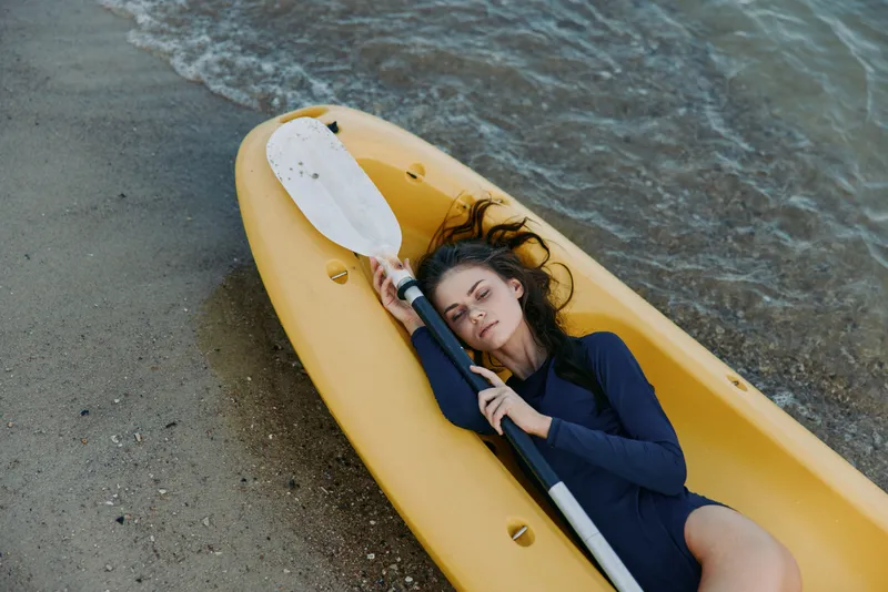 A person lies inside a yellow kayak on a sandy beach, eyes closed, resting while gentle waves lap nearby, capturing a peaceful moment of travel and leisure.