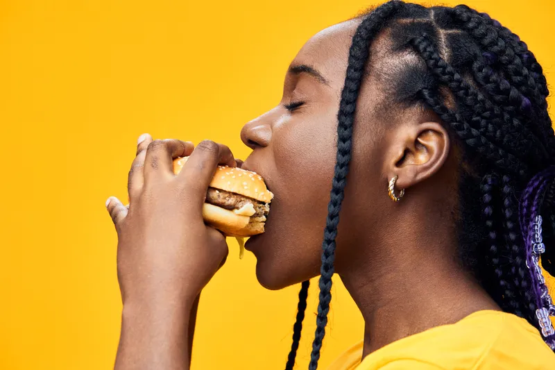 Young Black woman enjoying a hamburger with a bright yellow background, showcasing happiness and the joy of indulgent eating, perfect for food and lifestyle themes