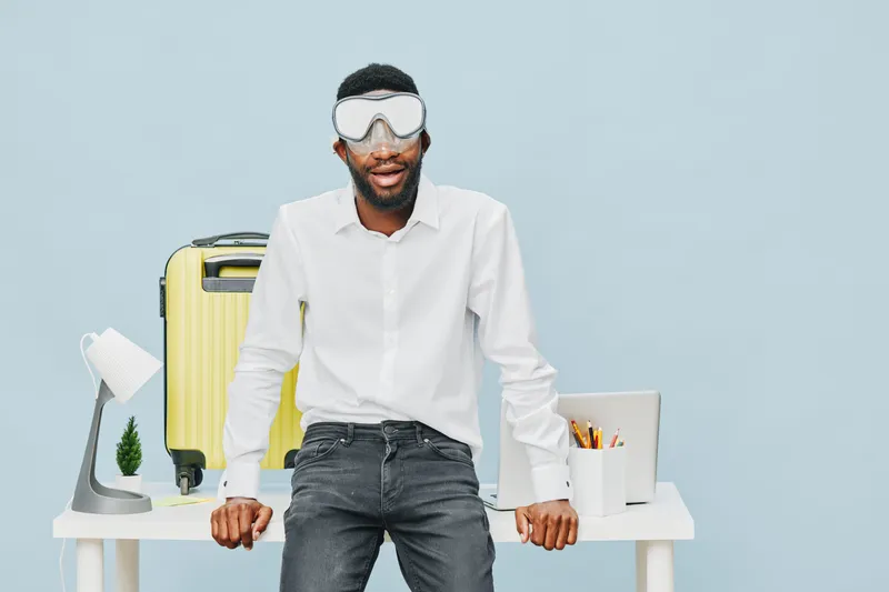 Modern young man with beard wearing sleep mask, casual white shirt and jeans, standing confidently at home office desk with travel luggage, laptop and stationery, concept of remote work and travel rel