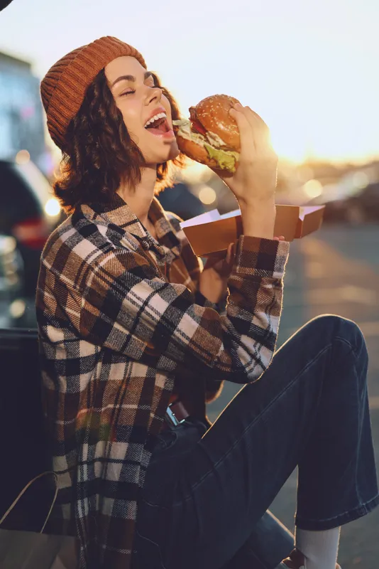 Woman eating burger with laughter in candid urban moment, authenticity and lifestyle vibe at golden hour glow. Emotional storytelling, mindful living and modern street aesthetic.
