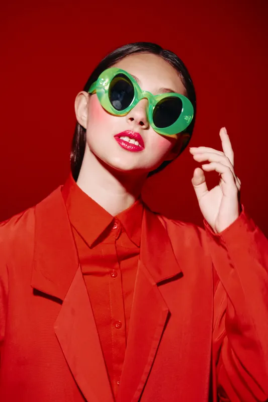 Fashion portrait of confident young woman wearing vibrant green sunglasses and bold red lipstick in focus against a red background, showcasing creative beauty and style.
