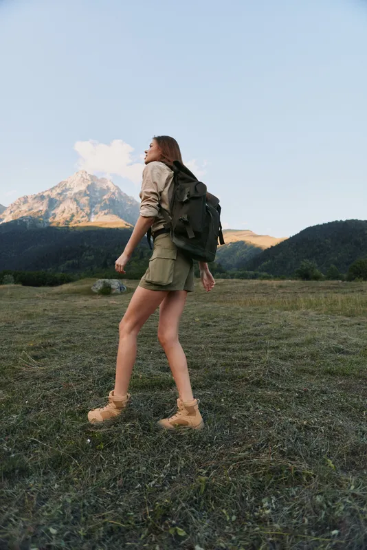 Young woman with long hair hikes across a grassy field with a large backpack under a clear sky and mountain landscape in the background.