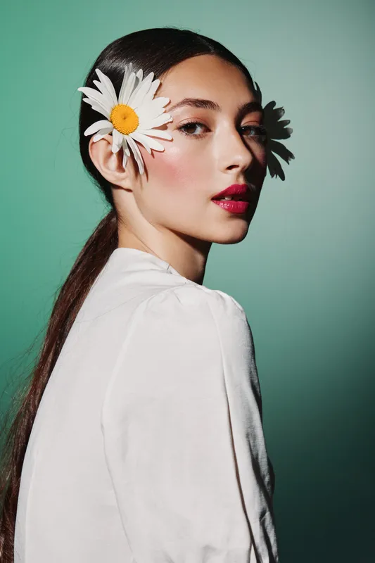Portrait of a young woman with daisy flowers in her hair wearing bold makeup and a white top. Studio shot with teal background for beauty, fashion, and creative concept.