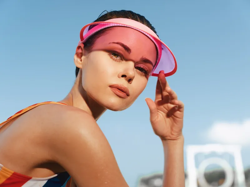 Close-up portrait of an athletic woman outdoors under a clear blue sky, wearing a pink visor and swimsuit, sunlit skin, confident gaze, and vibrant sports vibe.