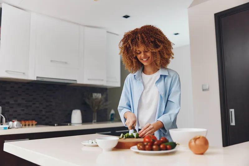 Счастливая афроамериканка готовит полезный салат на домашней кухне, используя свежие ингредиенты и кухонную утварь