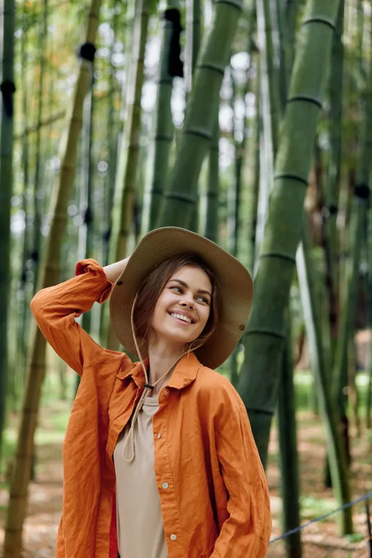 Woman in hat and orange jacket smiling in bamboo forest portrait, happy traveler enjoying nature with soft sunlight, vertical composition, relaxed outdoor fashion and warm tones.