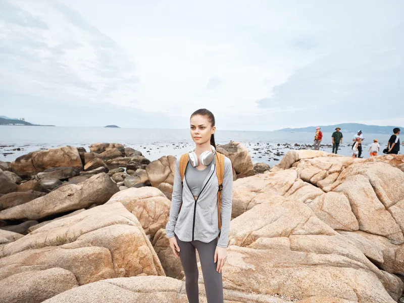 Young woman in activewear with headphones standing on rocky beach, reflecting a relaxed and focused lifestyle on a cloudy day