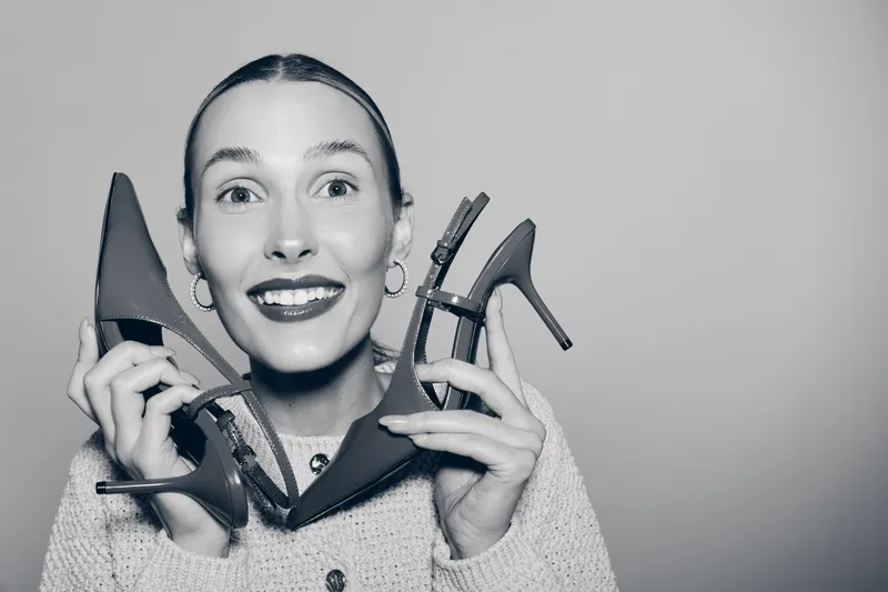 Happy white woman with fair skin holds high heel shoes, smiling with teeth visible, isolated on plain background.