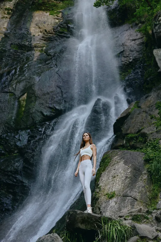 young woman standing near waterfall in activewear, surrounded by lush greenery and rocky cliffs, embodying nature and fitness harmony in outdoor setting