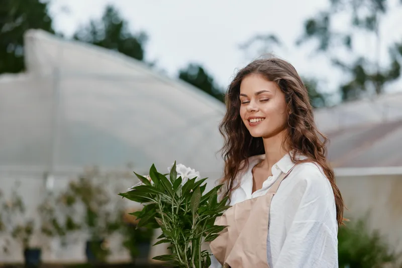 Young woman in white shirt and apron holding flowers, smiling in a serene garden setting with soft colors, showcasing joy and a connection to nature