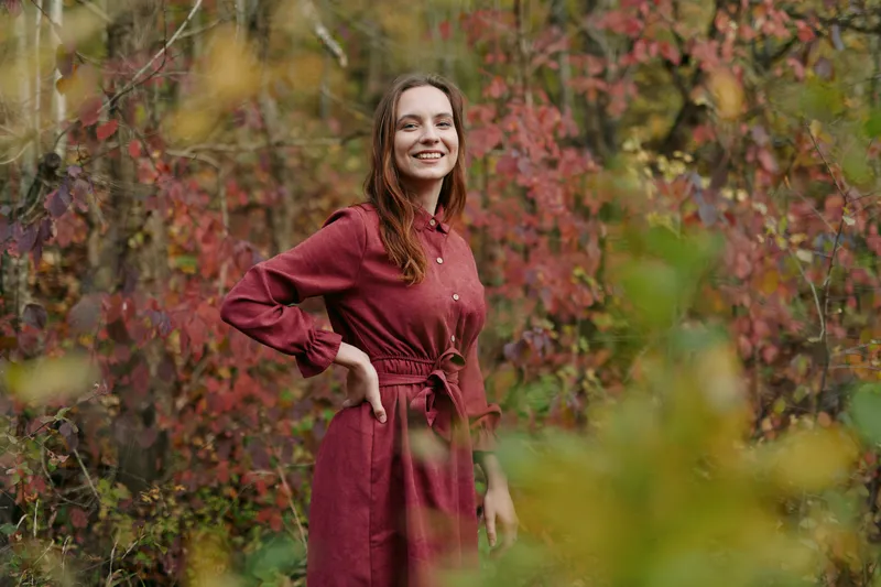 A portrait of a smiling woman in a burgundy dress in a autumn forest, capturing authenticity and credibility through natural light and a relaxed, genuine pose.