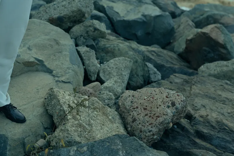 Close up of rugged rocks along a shoreline with a formal shoe and pant leg stepping onto stone, highlighting natural textures and a calm coastal atmosphere in muted light