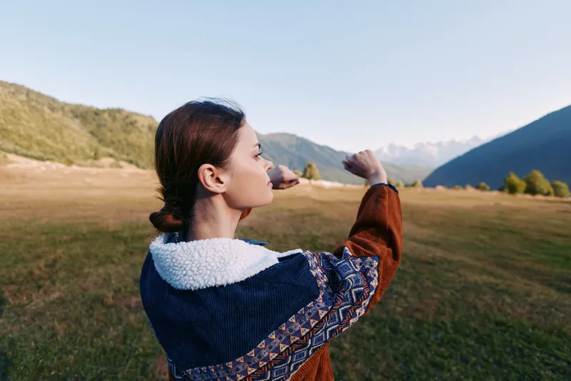 Woman in colorful jacket standing in a mountain meadow, profile view outdoors exploring nature, stretching arms toward sky in hiking pose, peaceful landscape and travel vibe