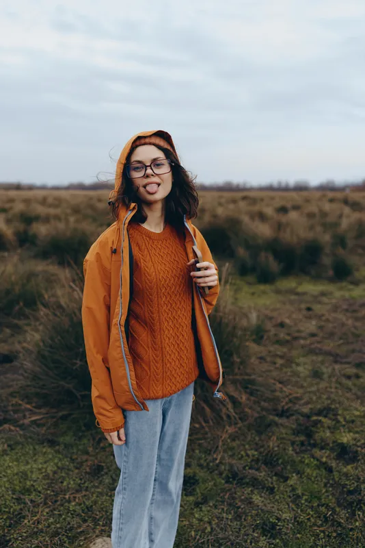 Woman standing outdoors in an autumn grassy field wearing an orange hoodie and glasses, casual lifestyle, soft overcast light, calm expression, nature background