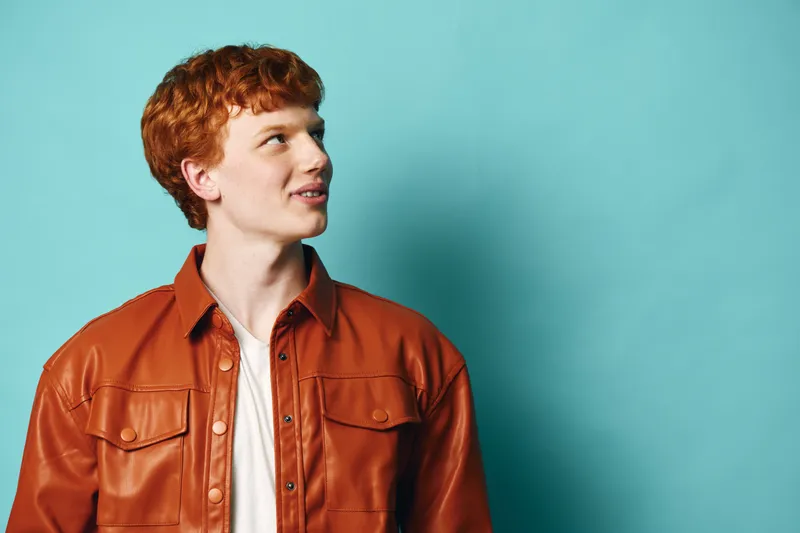 Young man with red hair wearing a brown leather jacket standing against blue background looking to the side. Casual fashion and portrait style with confident expression.