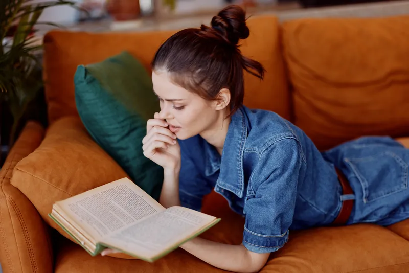 Женщина читает книгу, лёжа на животе на диване и опираясь головой на руки, в уютной гостиной