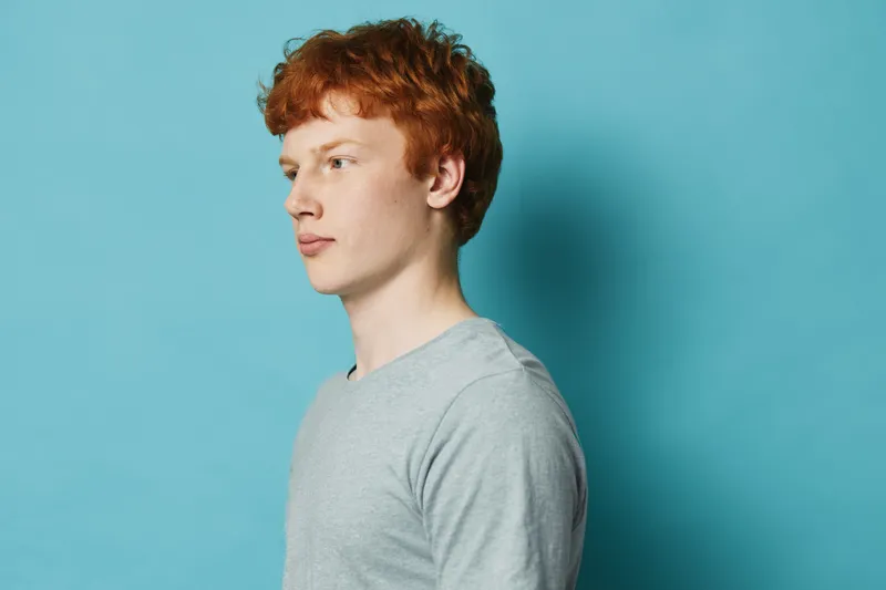 Young man with red hair and pale skin wearing grey sweater looking sideways on blue background. Portrait, casual style and natural light for studio shot and isolated concept.