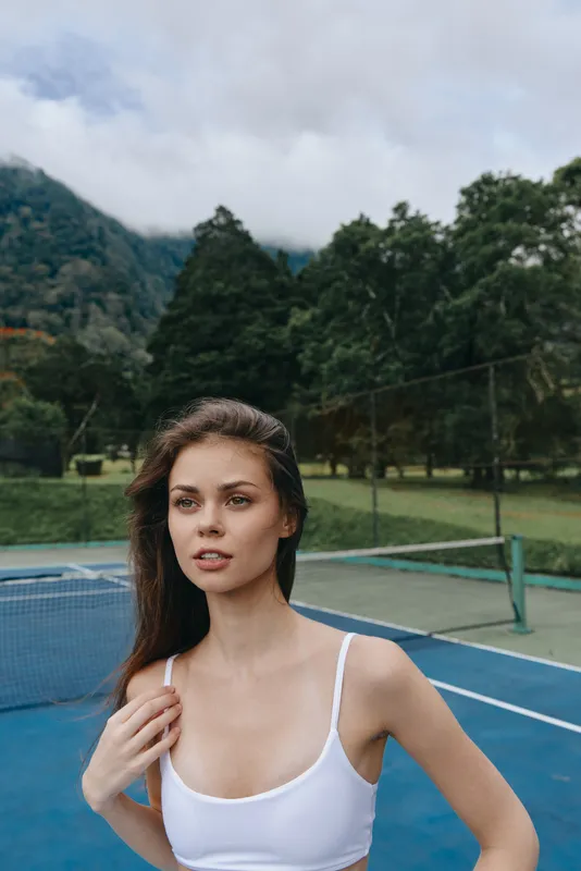 Serene young woman in a white sports bra on a tennis court, showcasing confidence and athleticism in a lush green setting with soft clouds in the background