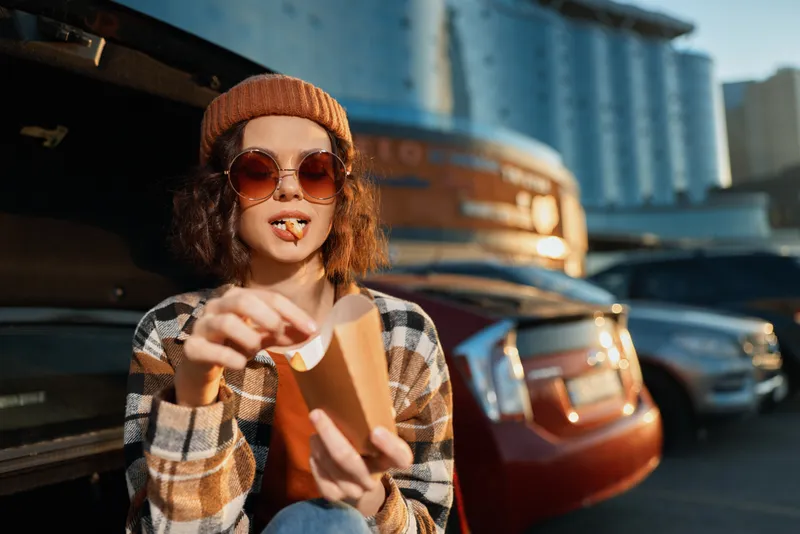 girl car fries parking sunglasses beanie trunk golden hour candid lifestyle portrait of a young woman eating fries by a car trunk, authenticity, mindful living and emotional storytelling in warm