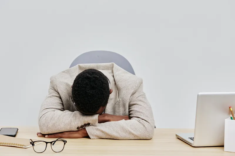 Black and white photo of a tired man resting his head on his arms at a desk with a laptop, glasses, and a smartphone, conveying fatigue or stress in a modern office environment. Business work concept.