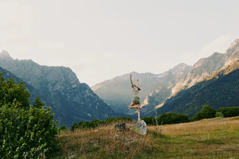 Young woman leaps on a rock in a sunlit alpine meadow with distant rugged mountains, radiating freedom and joy in an outdoorsy, carefree moment