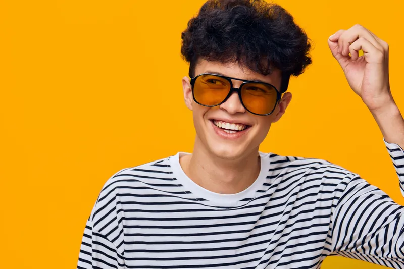 Smiling man wearing striped shirt and orange sunglasses enjoys vibrant yellow background, expressing happiness and youthful energy with curly hair and casual style.
