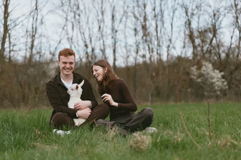 Goat, kid, couple outdoors in a meadow, grass, nature, happiness as they share a gentle moment with the animal, candid lifestyle scene, sunlit field, vibrant greens, relaxed connection