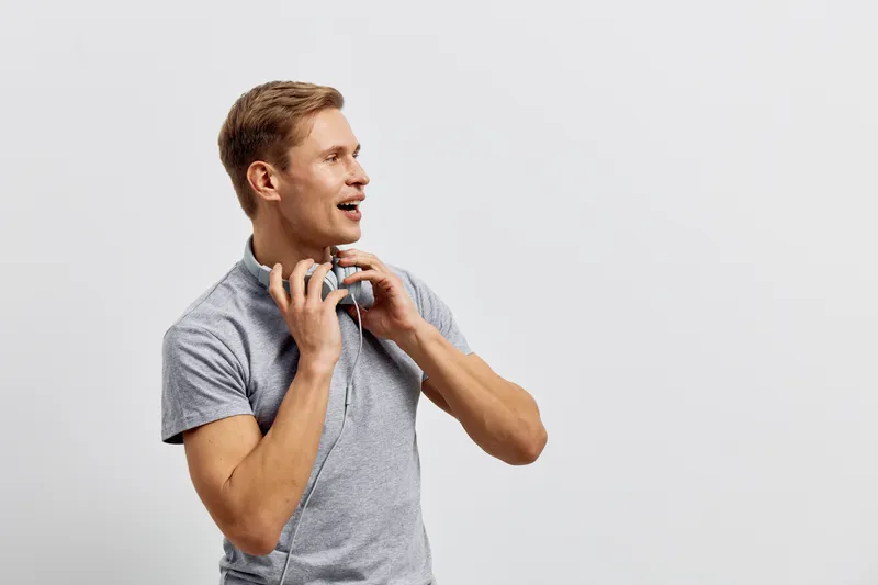 Man wearing casual gray t-shirt listening to music with headphones, expressing joy and excitement, standing against plain white background, studio portrait, lifestyle concept.