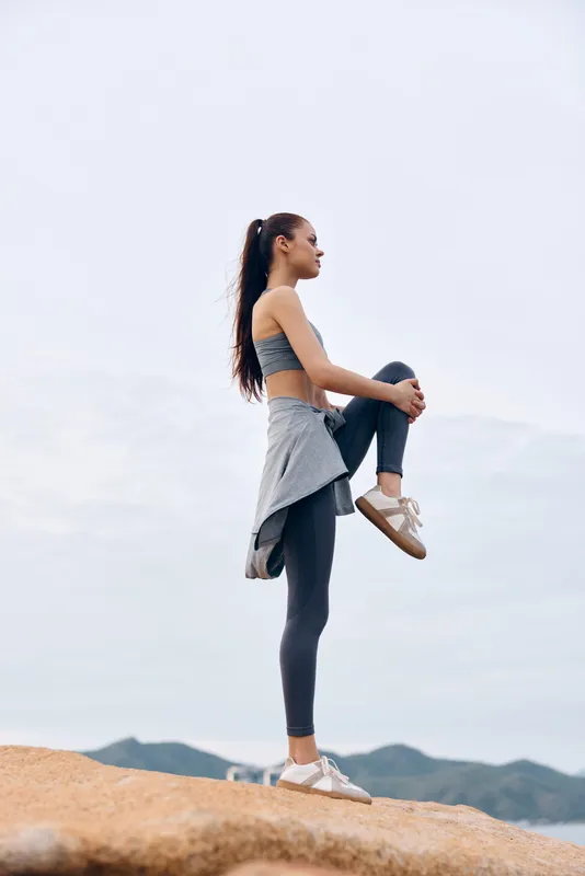 Young athletic woman stretching outdoors in sporty attire against a scenic backdrop, promoting fitness and healthy lifestyle concepts