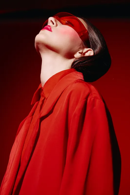 Fashion model wears red transparent glasses and a matching red coat, looking upwards in studio light. Creative beauty portrait, themes of style, color, and modern fashion.