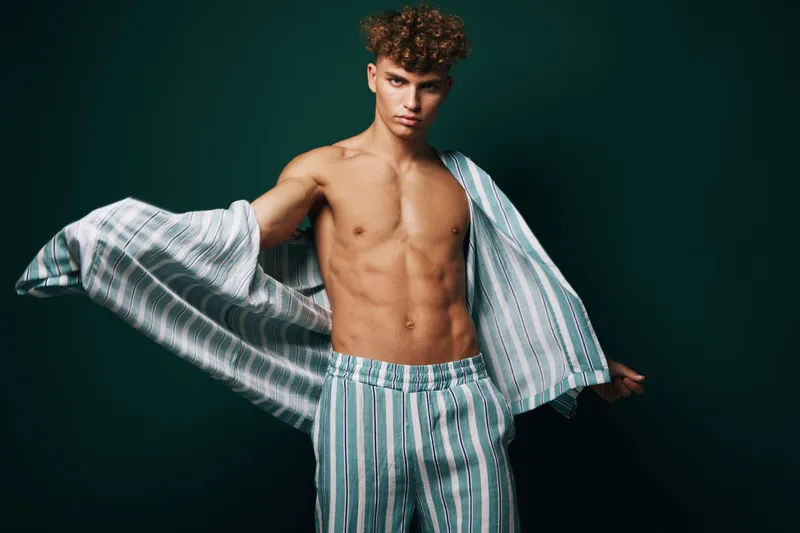 Young man with curly hair posing shirtless wearing striped pants and open shirt against dark background in studio for fashion and style concept.