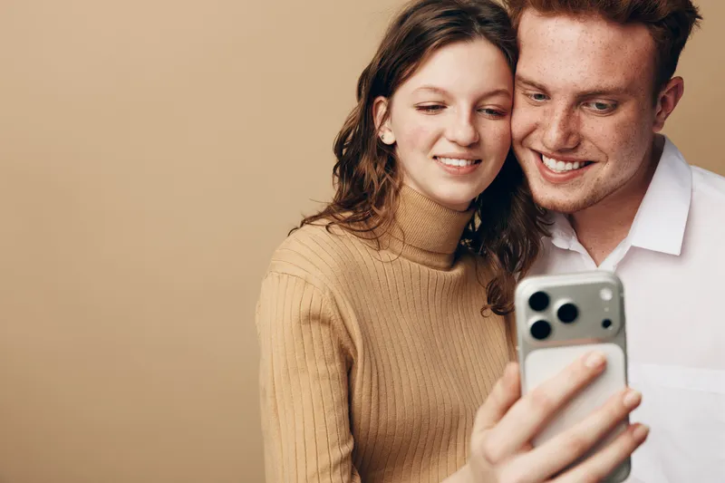 Young couple with happy expressions taking selfie on smartphone. Woman in beige sweater and man in white shirt smile together on beige background. Concept of love, technology, and friendship.