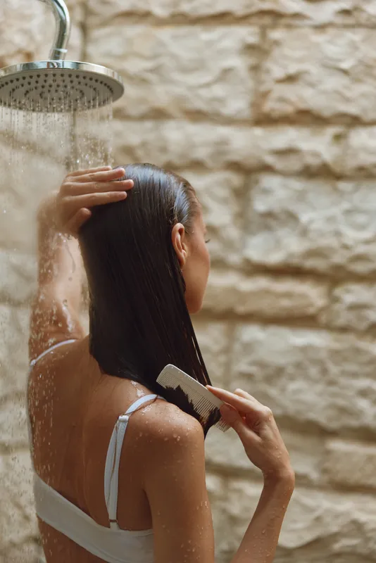 woman combing wet hair under shower with stone wall background, natural skin, relaxed posture, indoor bathroom setting, smooth dark hair strands