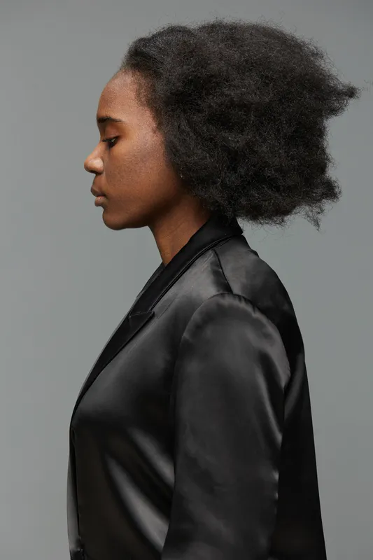 Profile portrait of a young Black woman with natural afro hair, wearing a sleek black blazer on a minimalist gray background, exuding confidence and elegance