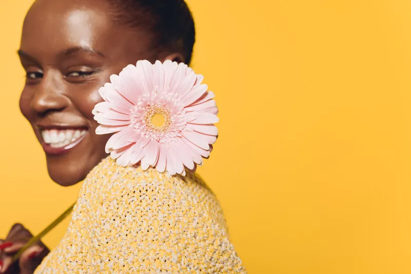 Smiling dark skinned African woman with short hair holds pink flower behind shoulder, wearing yellow sweater. Studio portrait on bright yellow background. Concept of happiness and natural beauty.