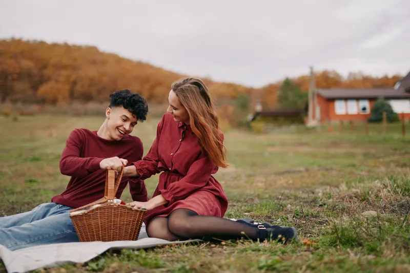 A young couple enjoys a genuine outdoor picnic on a grassy field, sharing laughter and opening a wicker basket together, captured in an authentic, candid moment of warm connection and ease.