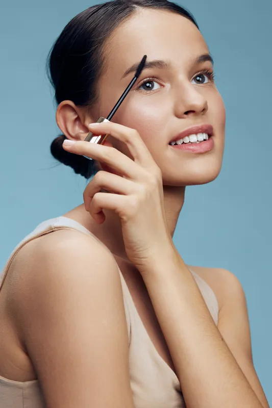 Young woman applying makeup, smiling with a brow pencil, showcasing a fresh look against a soft blue background, ideal for beauty and cosmetics promotions