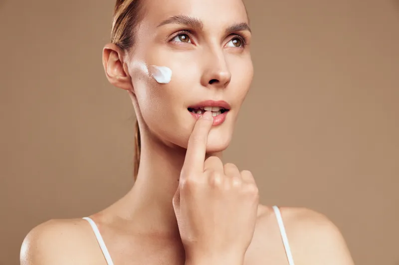 Woman applying skincare cream on cheek with finger, beauty routine closeup, smooth skin texture, calm expression, selfcare application, fresh, beige background