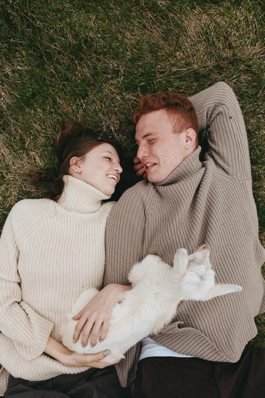 Joyful couple lying on grass outdoors, smiling at each other, soft sunlight, warm sweaters, relaxed pose, playful goat kid nearby, natural colors, countryside mood, cozy togetherness