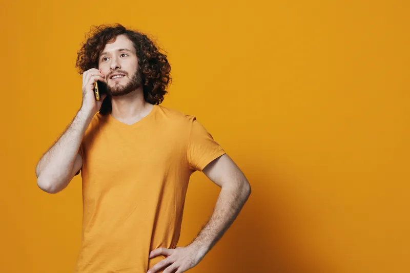 Cheerful young man talking on the phone, wearing an orange t shirt against a vibrant yellow background, expressing happiness and confidence