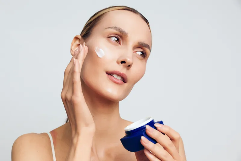 Woman applying skincare moisturizer to her face, using cream on cheek and holding a blue jar. Clean studio background shows daily beauty routine, skin care, selfcare.