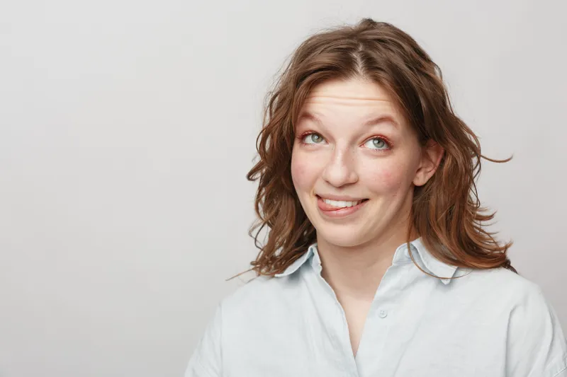 Young woman with wavy brown hair smiles with a playful expression wearing a white shirt on a light background. Concept of casual style, happiness, and natural beauty.