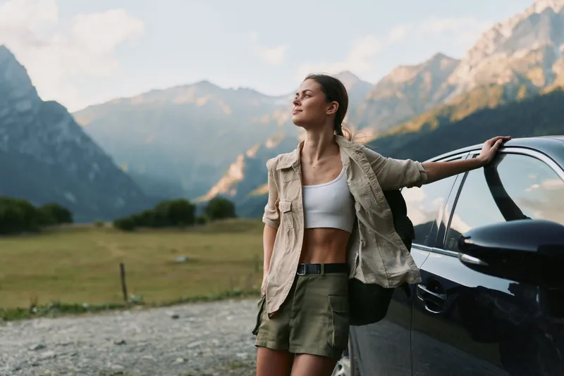 Young woman with long hair leans on a car door in a mountain valley during golden hour, enjoying a peaceful moment with a relaxed and confident expression.