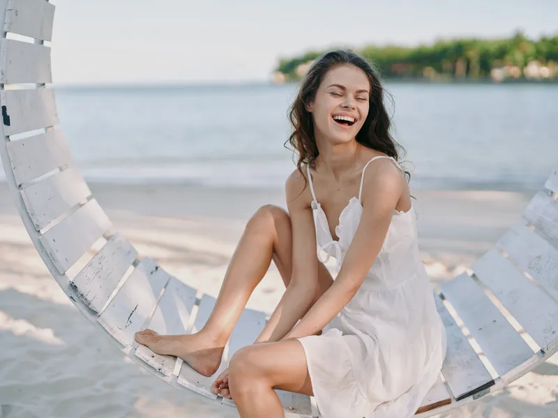 Young woman sits on a round white beach swing in a light white dress, laughing as waves wash the shore under a clear blue sky, capturing relaxed coastal fashion and playful joy.