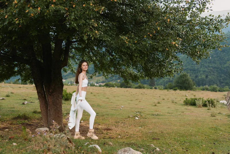 A young woman in white pants and a white top stands beside a large tree in an open field, smiling softly as she looks back, radiating carefree outdoor vibes.