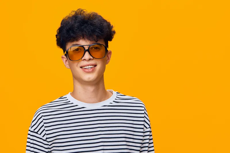 young man wearing striped shirt and yellow sunglasses smiling confidently on bright orange background with casual hairstyle and joyful expression